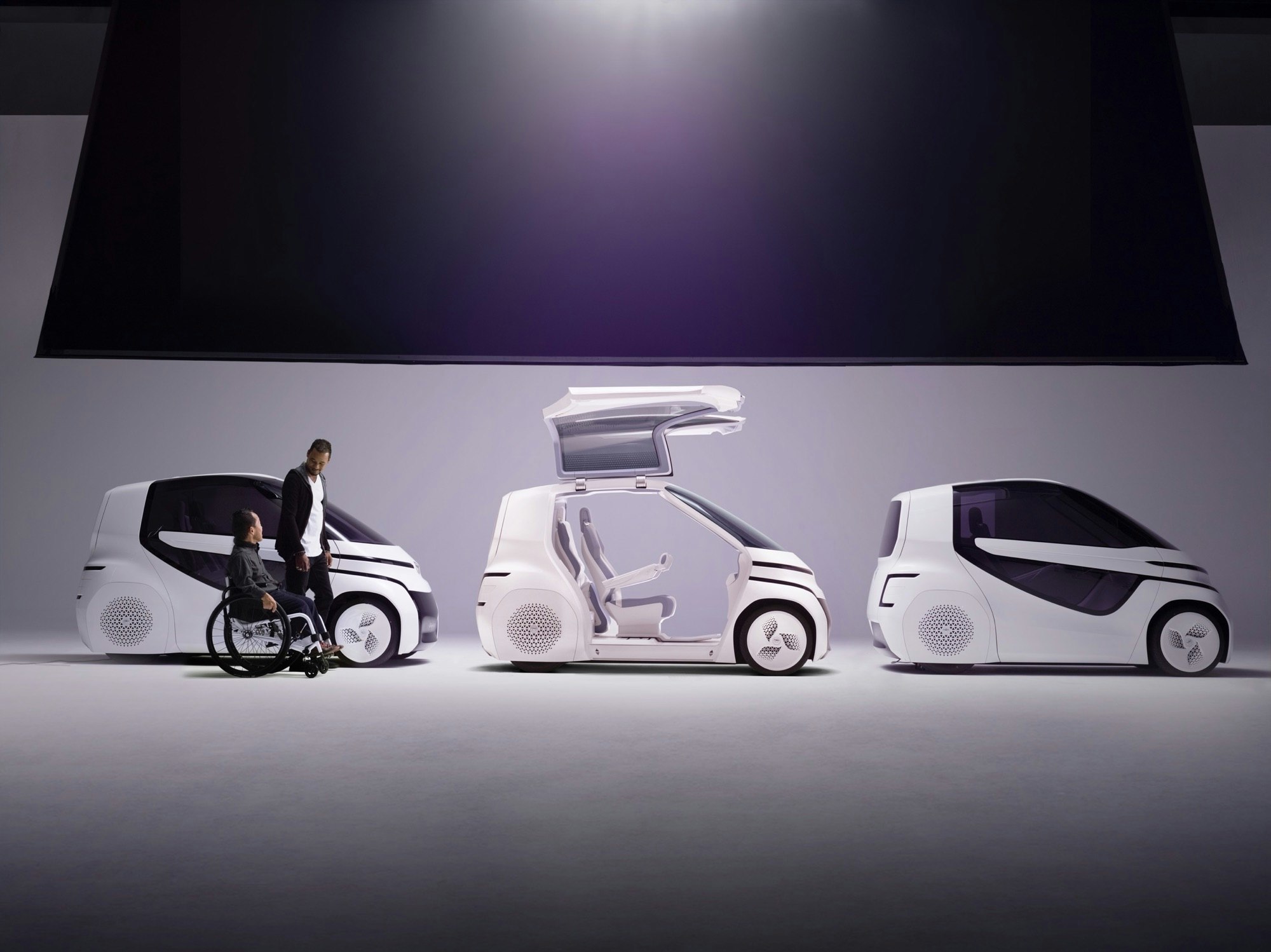 Three futuristic white automotive vehicle designs displayed in a studio, with one featuring a man and a person in a wheelchair.
