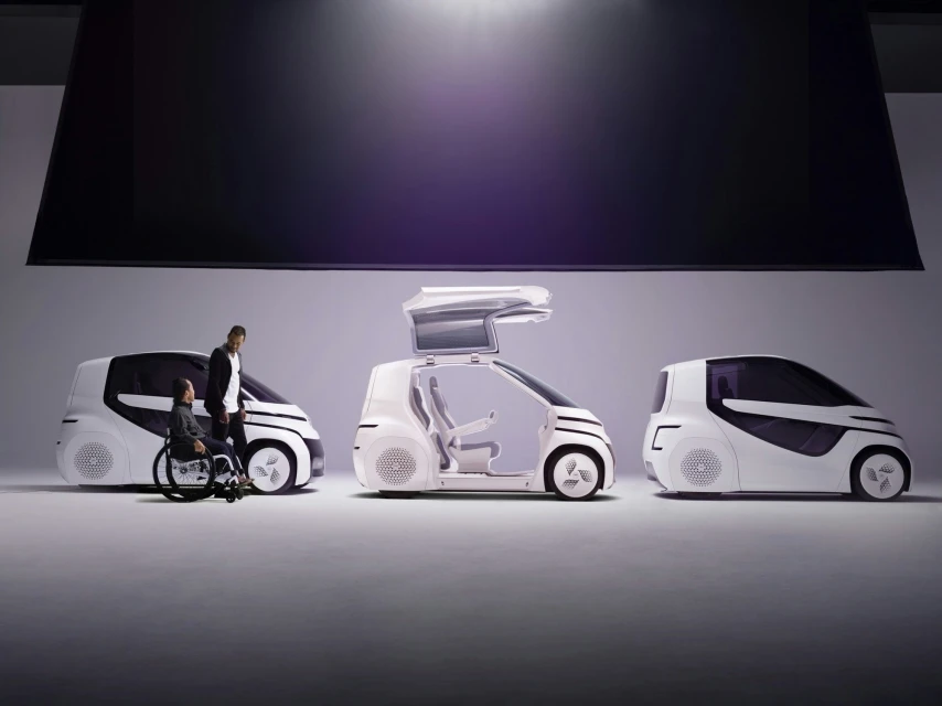 Three futuristic white automotive vehicle designs displayed in a studio, with one featuring a man and a person in a wheelchair.