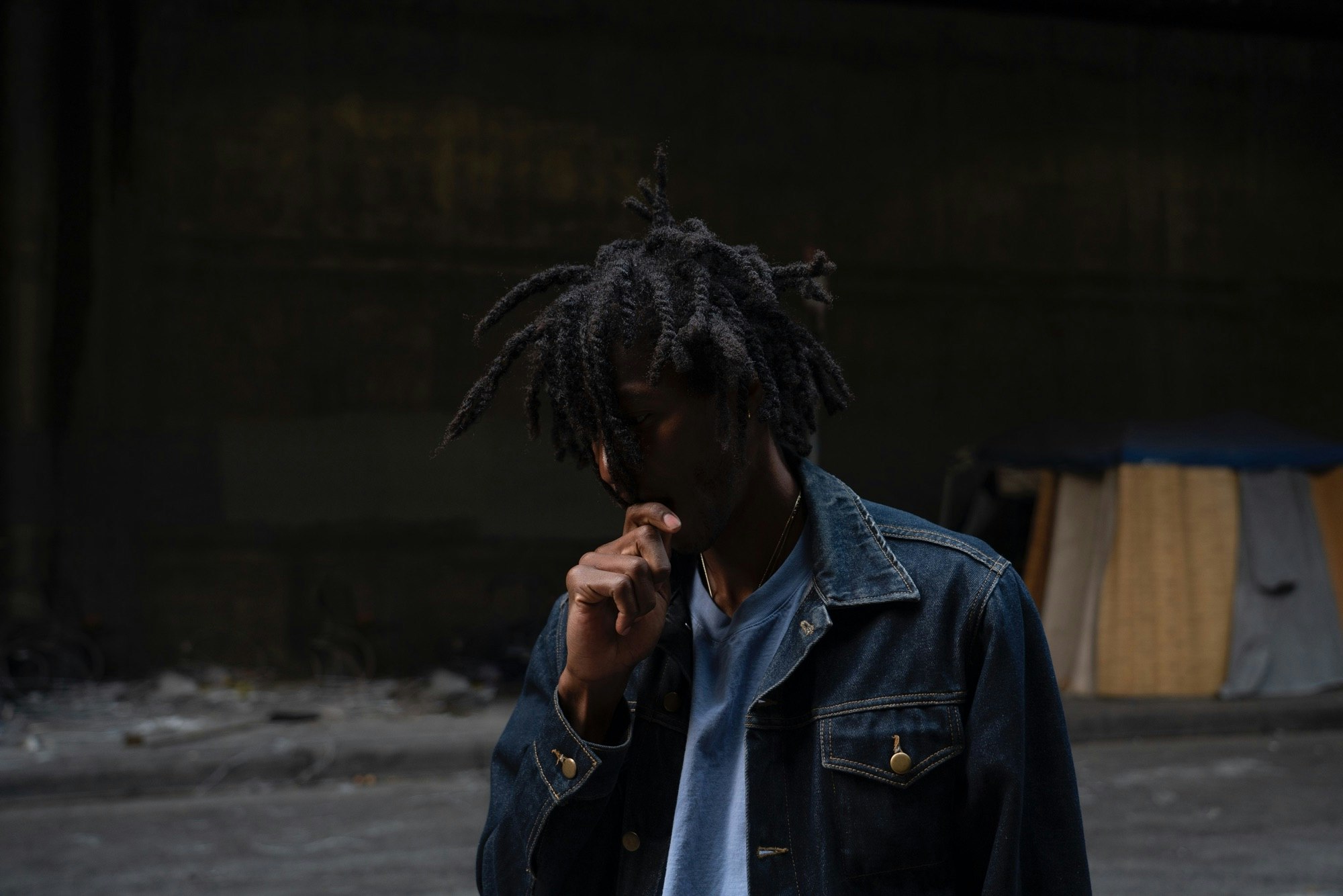 Adult male with dreadlocks, wearing a denim jacket, standing in an urban environment with a concrete floor and dark background.