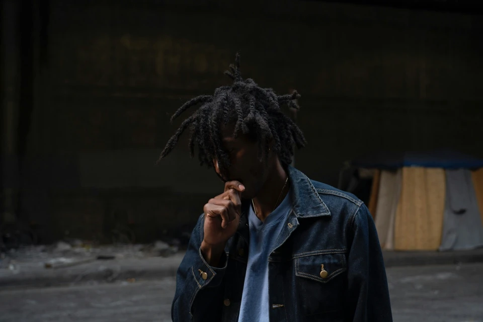 Adult male with dreadlocks, wearing a denim jacket, standing in an urban environment with a concrete floor and dark background.