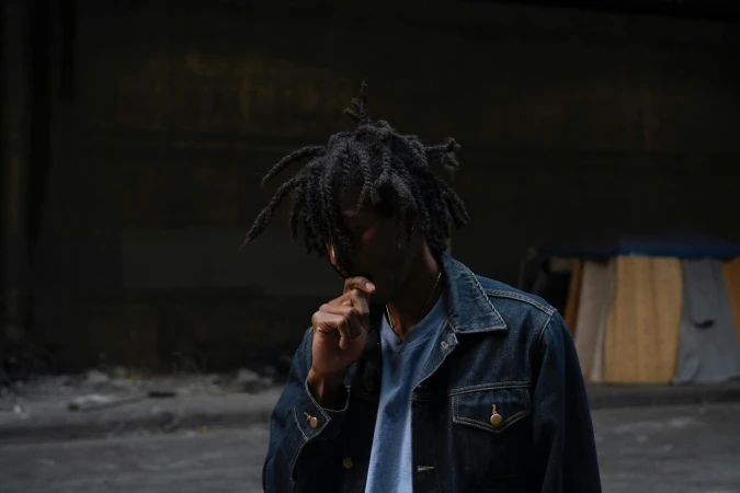 Adult male with dreadlocks, wearing a denim jacket, standing in an urban environment with a concrete floor and dark background.