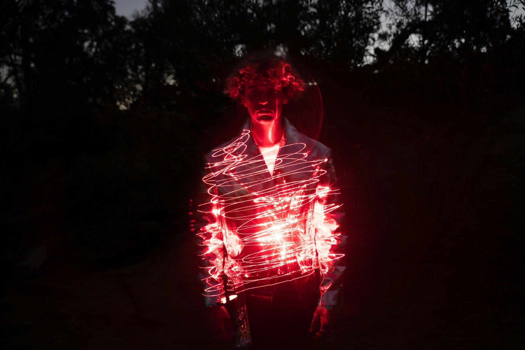 Person in jacket poses at night with light painting effect. Red glowing lines curve over their torso against a dark, wooded backdrop.