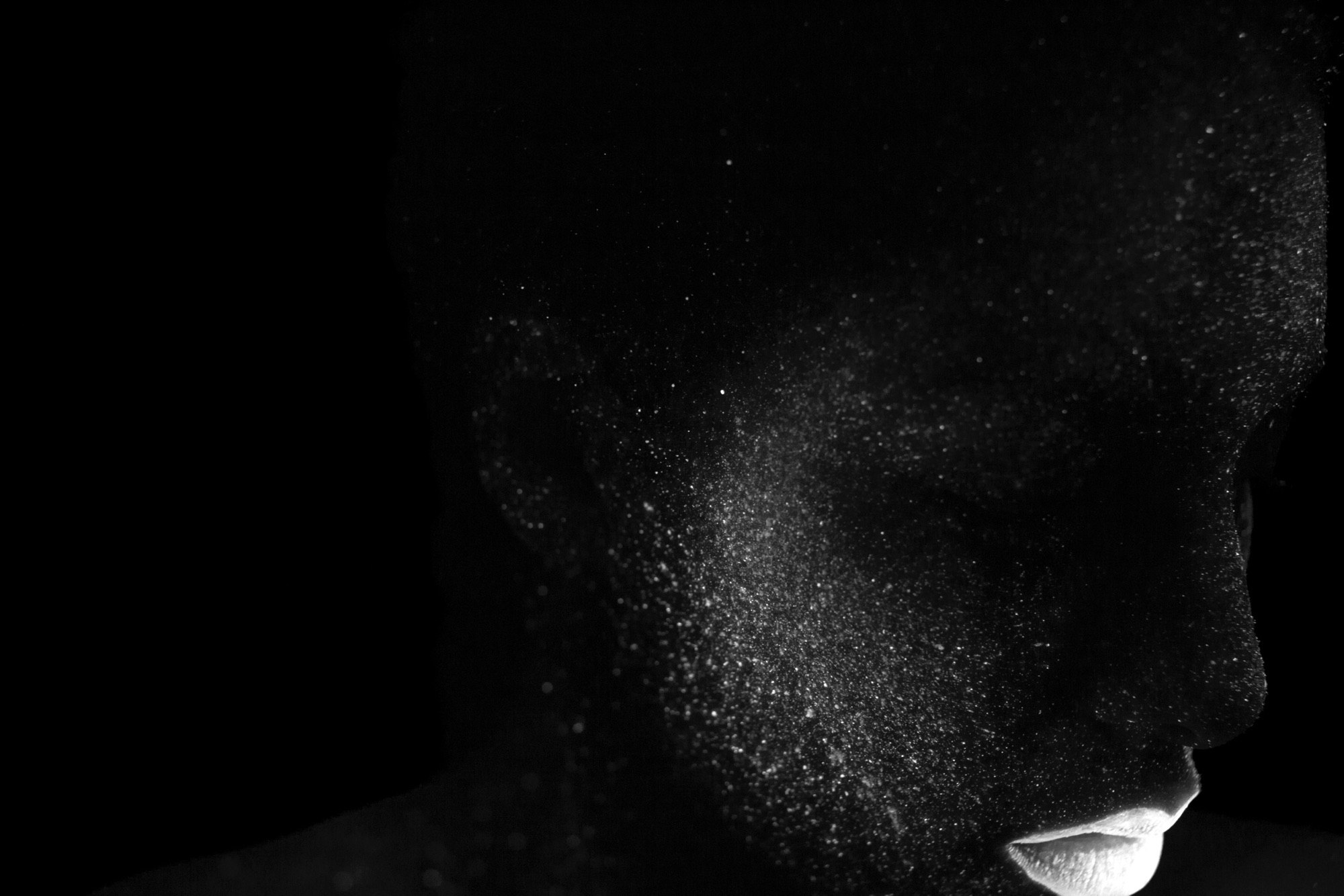 Close-up studio portrait of a person with dark skin covered in shimmering white powder against a black background, emphasizing texture and form.