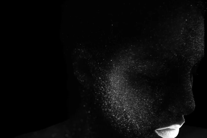 Close-up studio portrait of a person with dark skin covered in shimmering white powder against a black background, emphasizing texture and form.