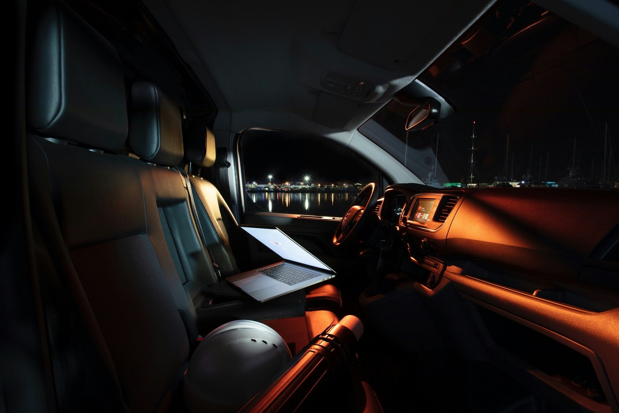 Interior view of a van at night with a laptop on the passenger seat, helmet, and a view of harbor lights in the background.