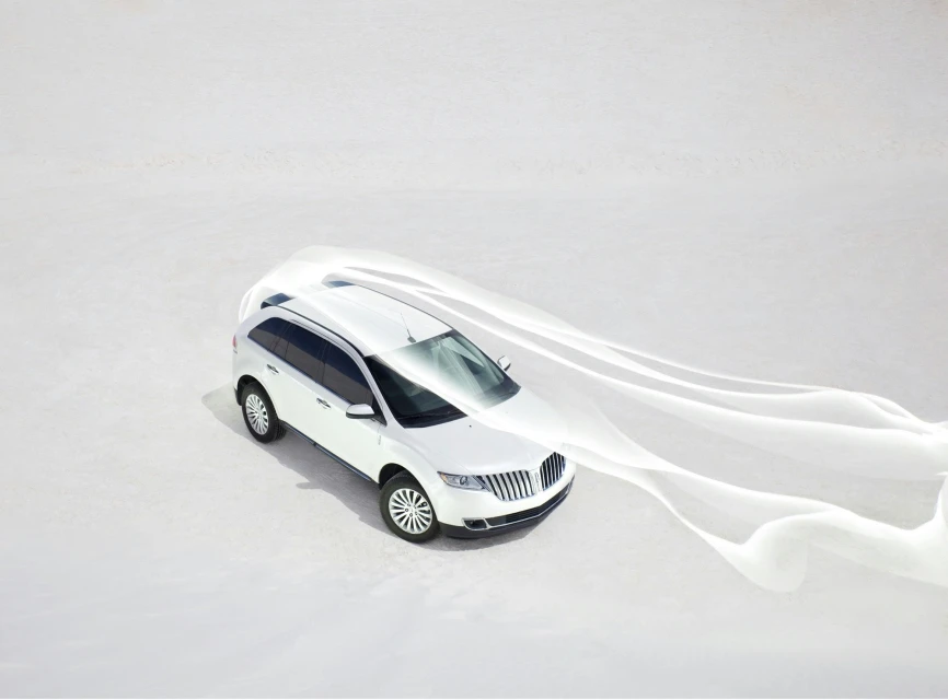 Aerial view of a white Lincoln SUV on a white surface, trailing white fabric, in bright daylight. Automotive and outdoor lifestyle imagery.