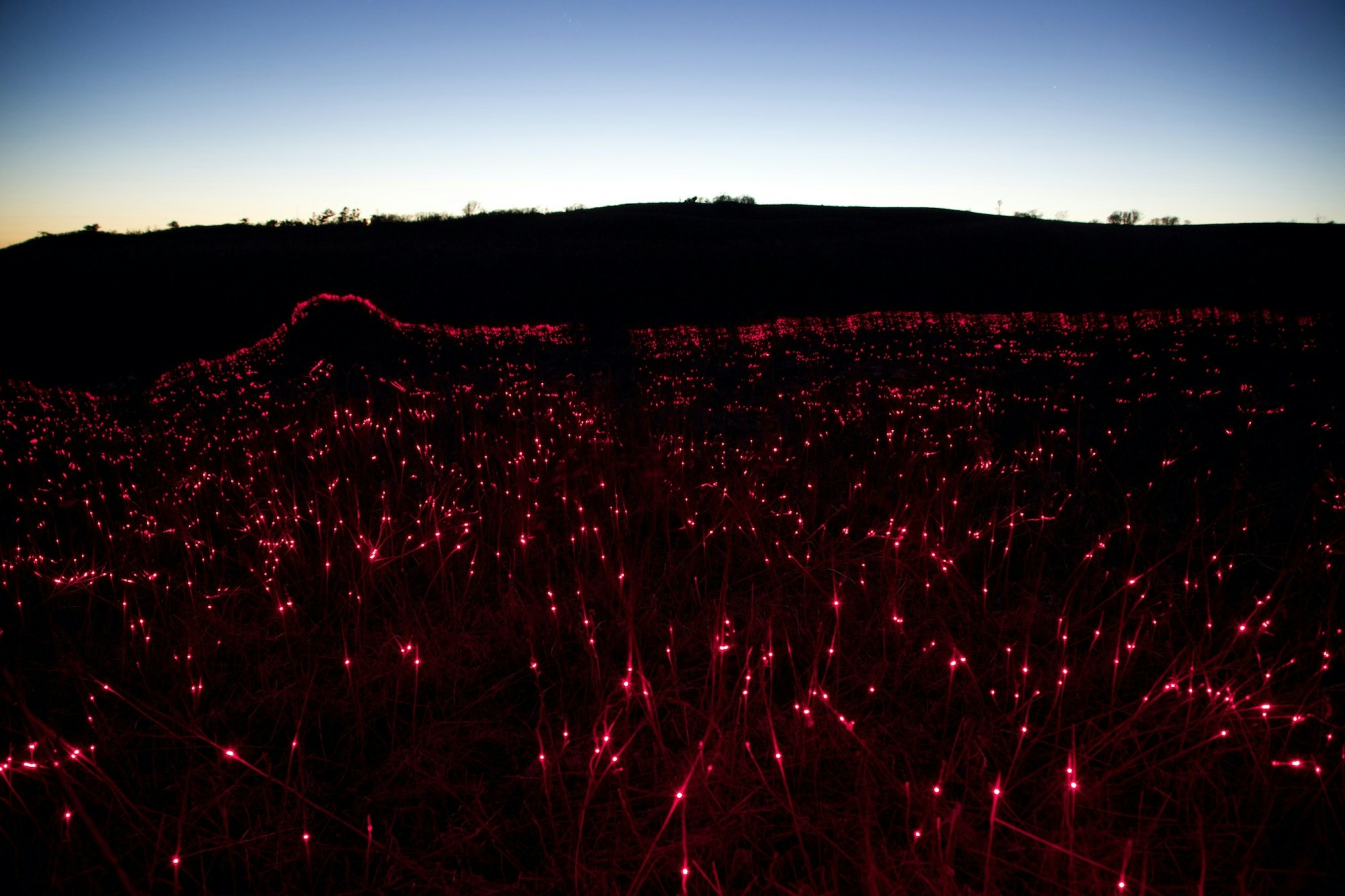 Red lights illuminate a field of dormant grass at dusk, silhouetted by a dark ridgeline against a gradient blue and orange sky.