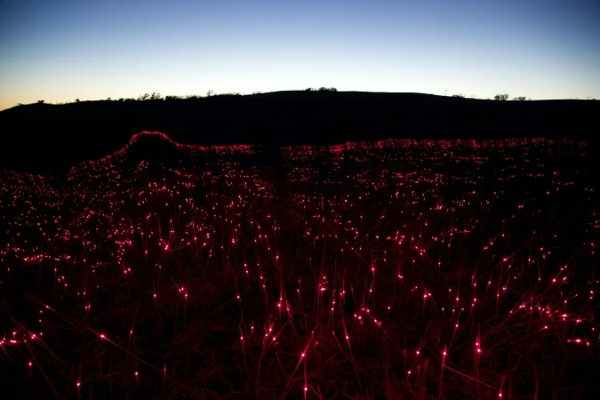Red lights illuminate a field of dormant grass at dusk, silhouetted by a dark ridgeline against a gradient blue and orange sky.