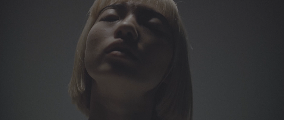 Low angle closeup portrait of a woman with short blonde hair and closed eyes against a neutral dark grey background.