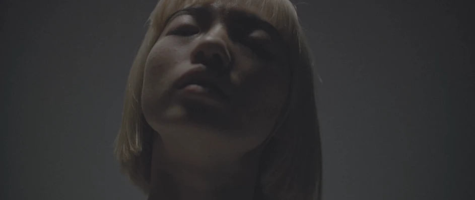 Low angle closeup portrait of a woman with short blonde hair and closed eyes against a neutral dark grey background.
