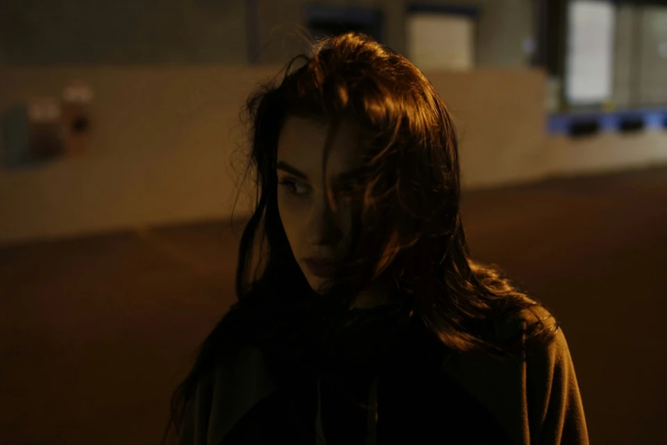 Close-up portrait of a young adult woman with long brown hair, looking off to the side in a dimly lit, urban setting at night.