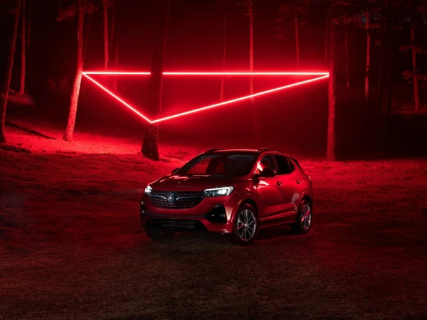 Red Buick SUV parked at night in forest with neon triangle art installation illuminated, creating a moody automotive scene.