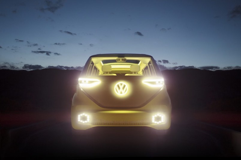Volkswagen ID Buzz electric van with headlights and logo illuminated at dusk against mountain range and night sky background.