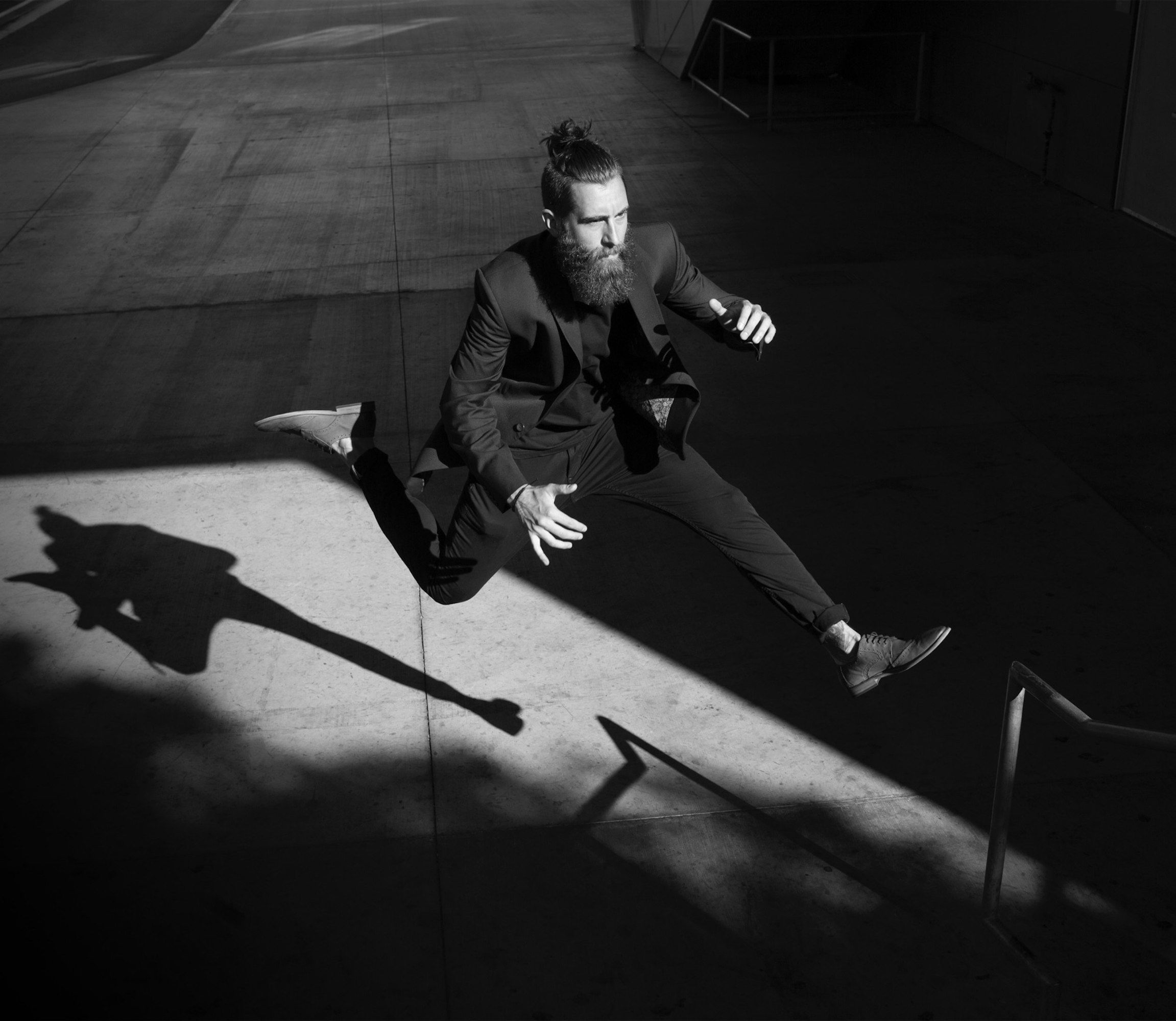 Black and white portrait of a bearded man in a dark suit jumping in the air with his shadow visible on the concrete surface.