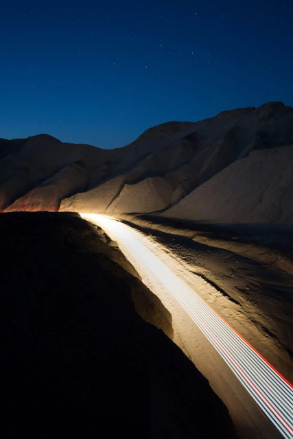 Nighttime desert landscape featuring light trails along a road with mountains under a dark blue sky and stars, creating a unique visual effect.