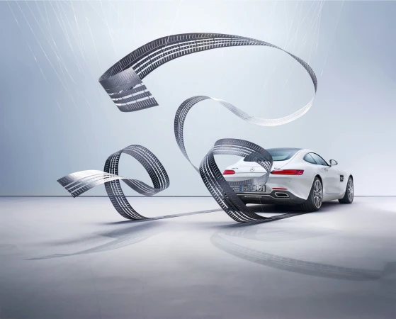 White Mercedes-AMG GT automobile is presented in a studio setting with a gray gradient backdrop and tire tread streamers.
