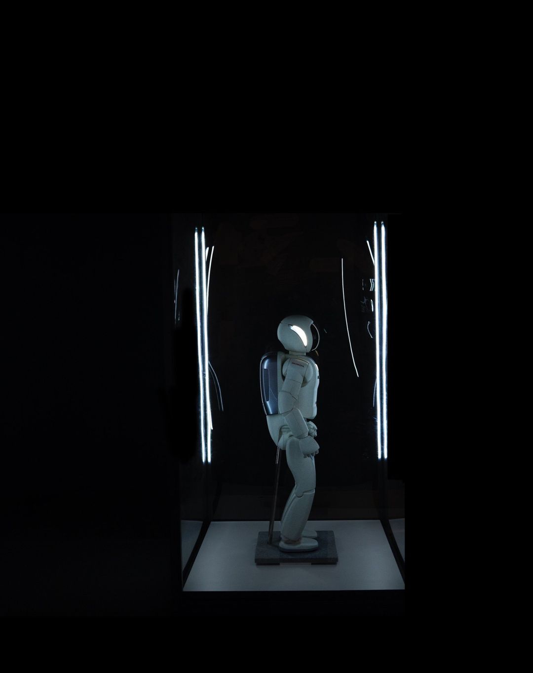 White robot stands under bright lights in a dark room. The robot appears to be on display in some kind of exhibit or showroom.
