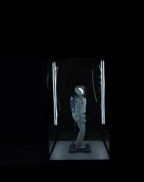 White robot stands under bright lights in a dark room. The robot appears to be on display in some kind of exhibit or showroom.