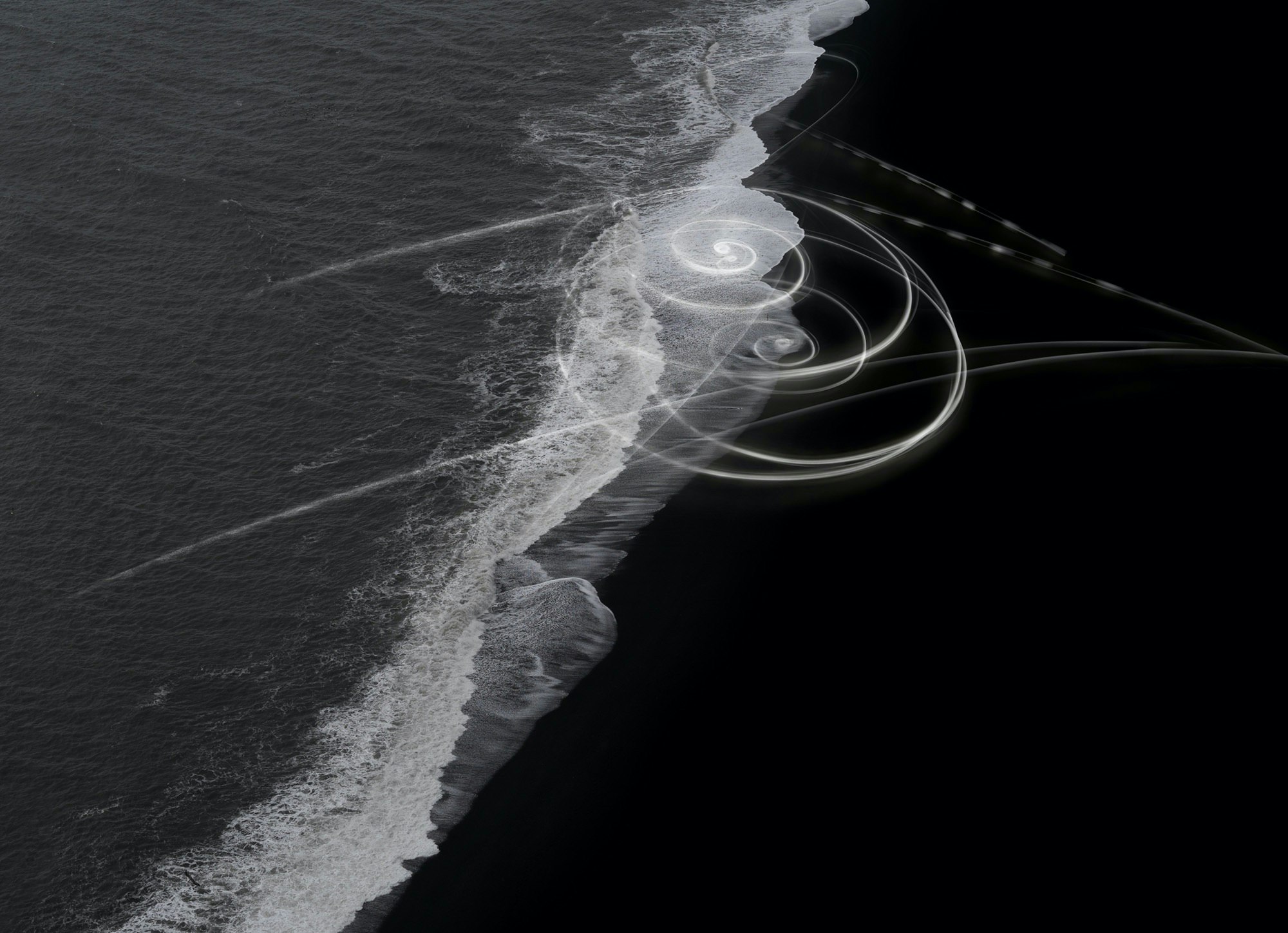 Abstract long exposure of ocean waves washing up on black sand beach with spirals of light painting adding surreal artistic touch.