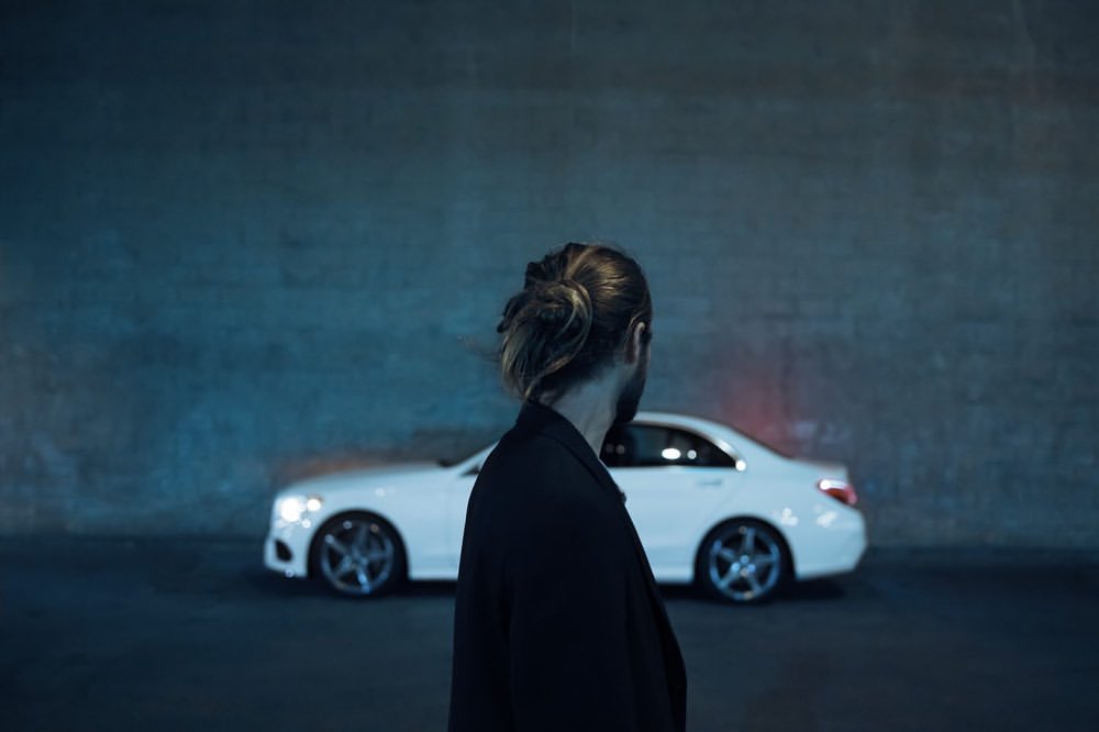 Adult male in a dark jacket faces away from the camera in front of a modern white car and a solid wall with diffused lighting.