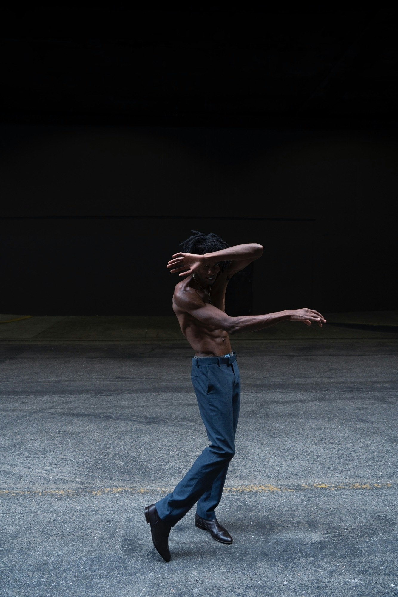 Man dancing with expressive pose in studio. He is shirtless with dark pants and dark shoes, expressive arm movement against dark background.