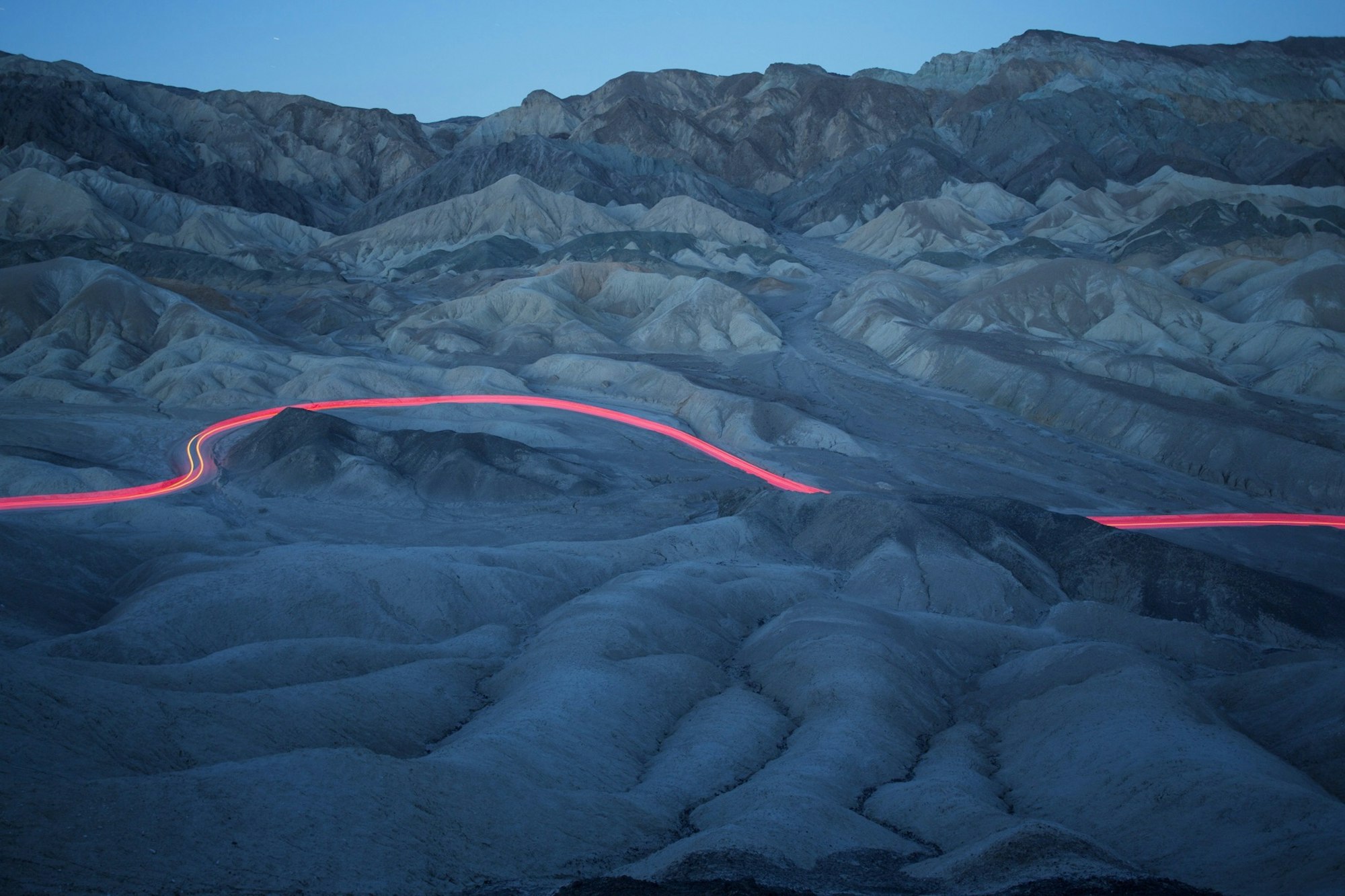 Mountains at dusk with a long exposure capturing light trails on a desert road, creating a dynamic contrast in a natural landscape.