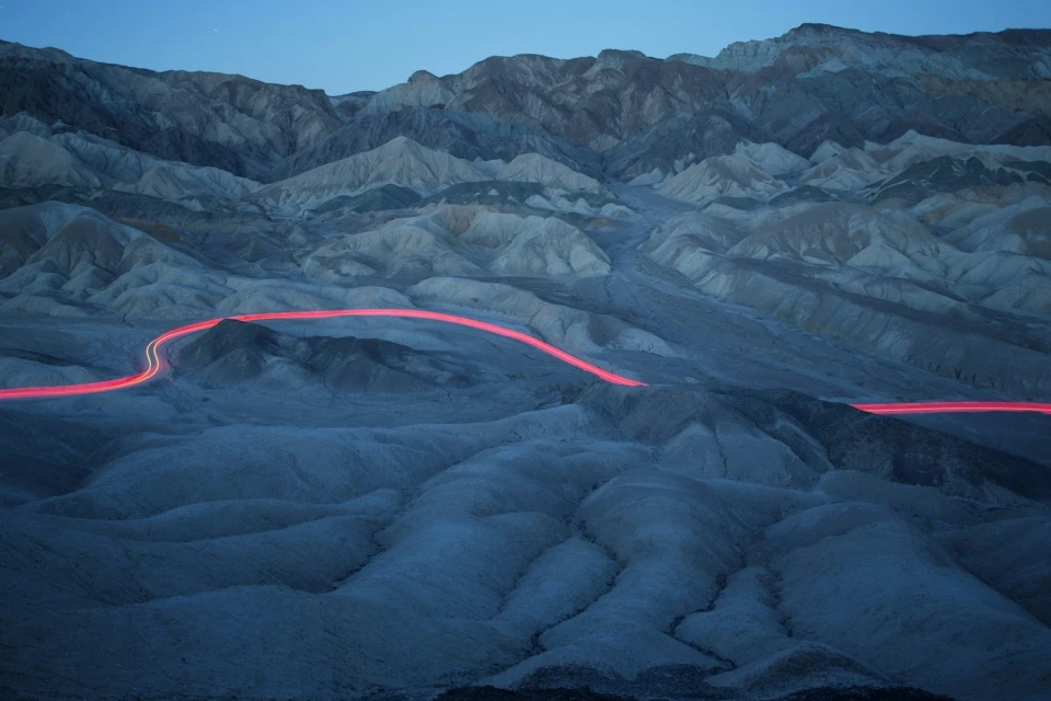 Mountains at dusk with a long exposure capturing light trails on a desert road, creating a dynamic contrast in a natural landscape.