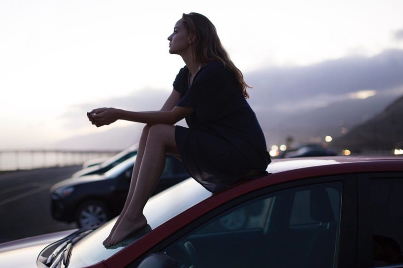 Woman in black dress sits barefoot atop a red car, overlooking a coastal highway at dusk with mountains and cloudy sky in the distance.