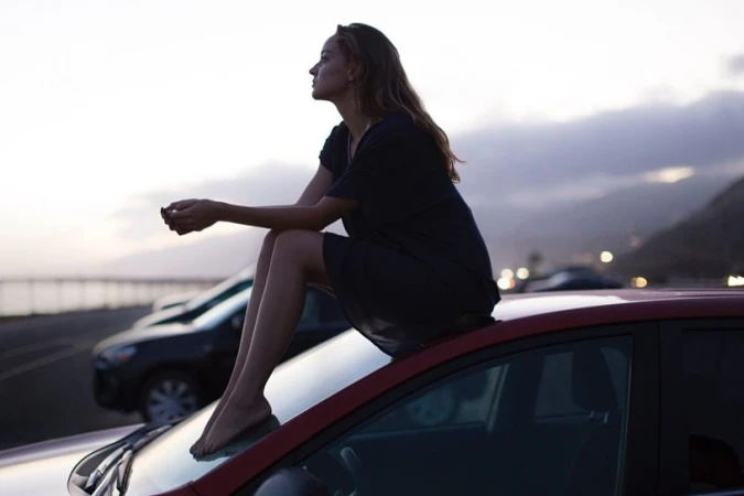 Woman in black dress sits barefoot atop a red car, overlooking a coastal highway at dusk with mountains and cloudy sky in the distance.