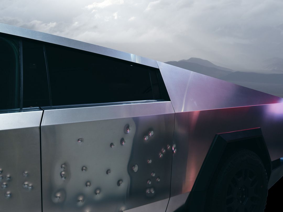 Side view of a metallic gray Tesla Cybertruck with simulated bullet holes, set against a blurred mountain landscape under a cloudy sky.
