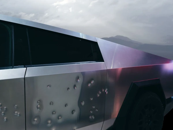 Side view of a metallic gray Tesla Cybertruck with simulated bullet holes, set against a blurred mountain landscape under a cloudy sky.