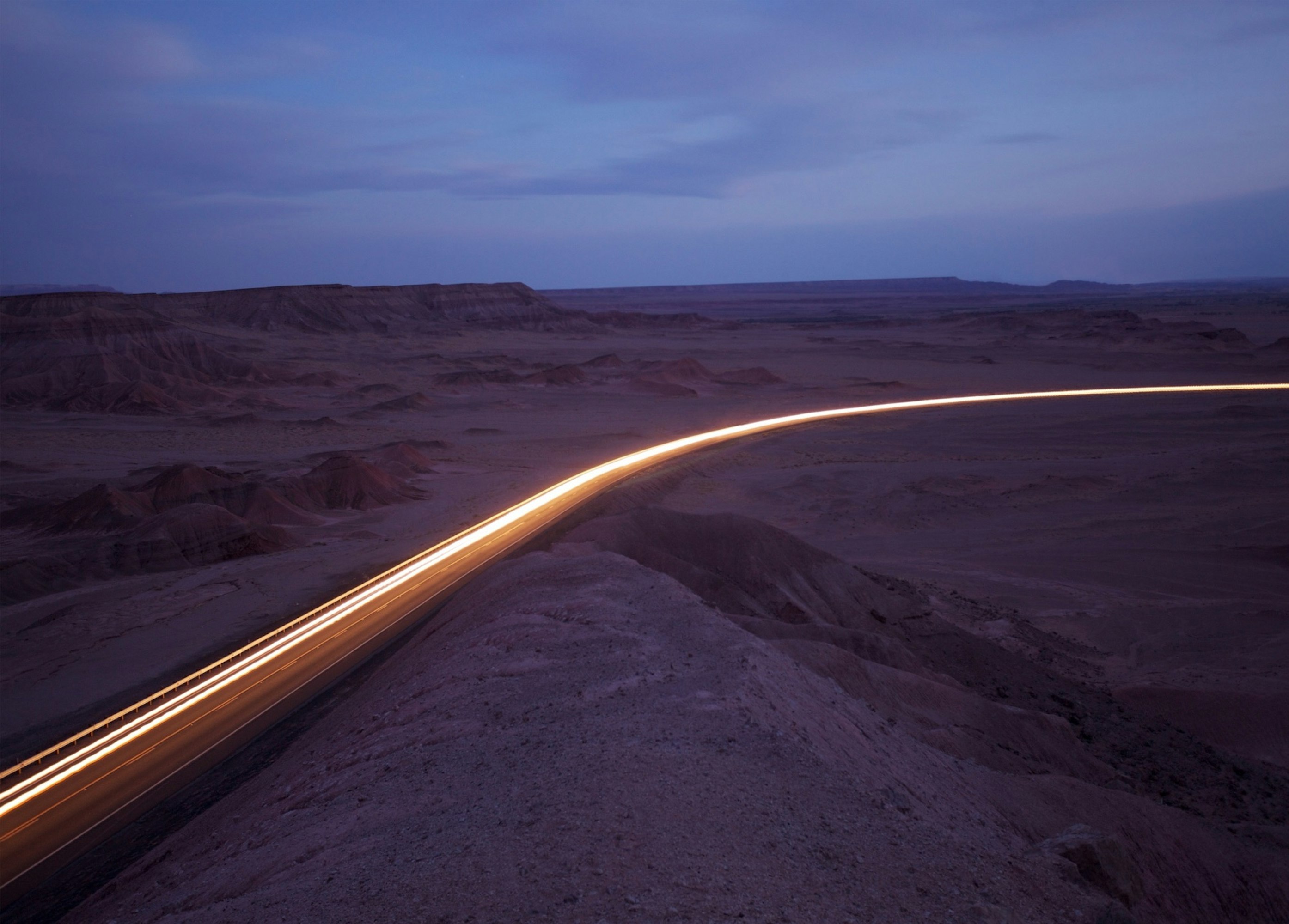Desert highway at night with light trails from passing cars under a blue sky, creating a sense of movement and solitude.