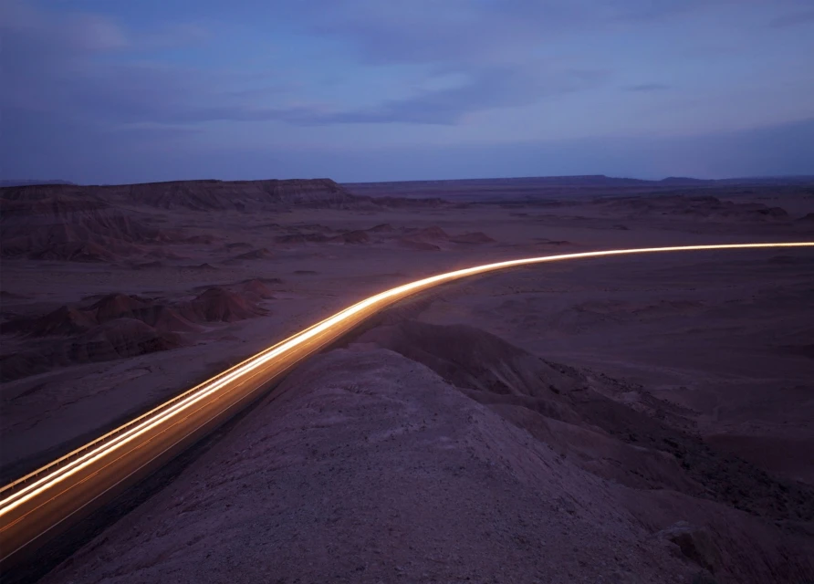 Desert highway at night with light trails from passing cars under a blue sky, creating a sense of movement and solitude.