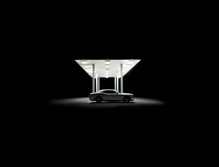 Silver coupe car parked under a brightly lit gas station canopy at night, set against a stark black background.