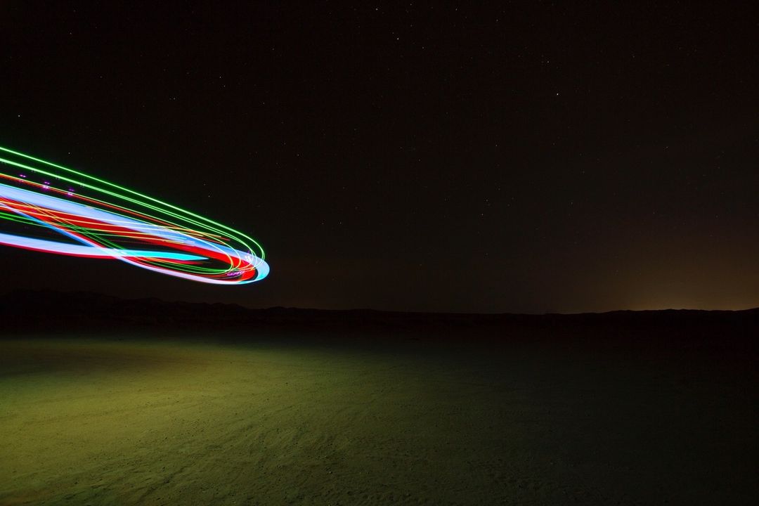Long exposure photo of light trails at night over dark, flat land under a starry sky with dark silhouette hills in the distance.