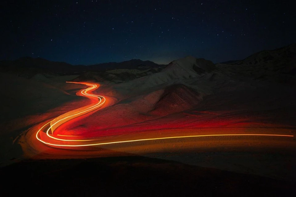 Long exposure landscape of a winding road cutting through a mountainous terrain at night under a star-filled sky with light trails.