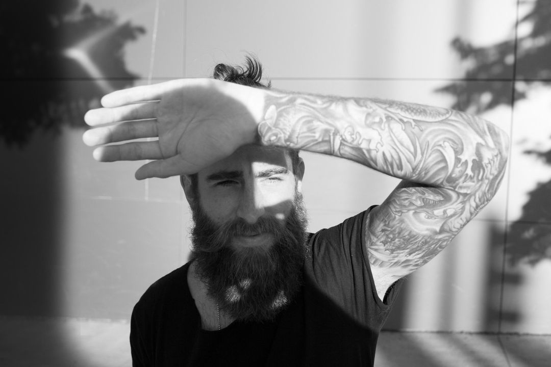 Black and white portrait of a bearded man with a top knot, shielding his eyes with his hand and displaying arm tattoos in outdoor light.