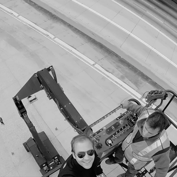 Two men operate an elevated work platform with controls, wearing safety vests, standing on a concrete surface with road markings.
