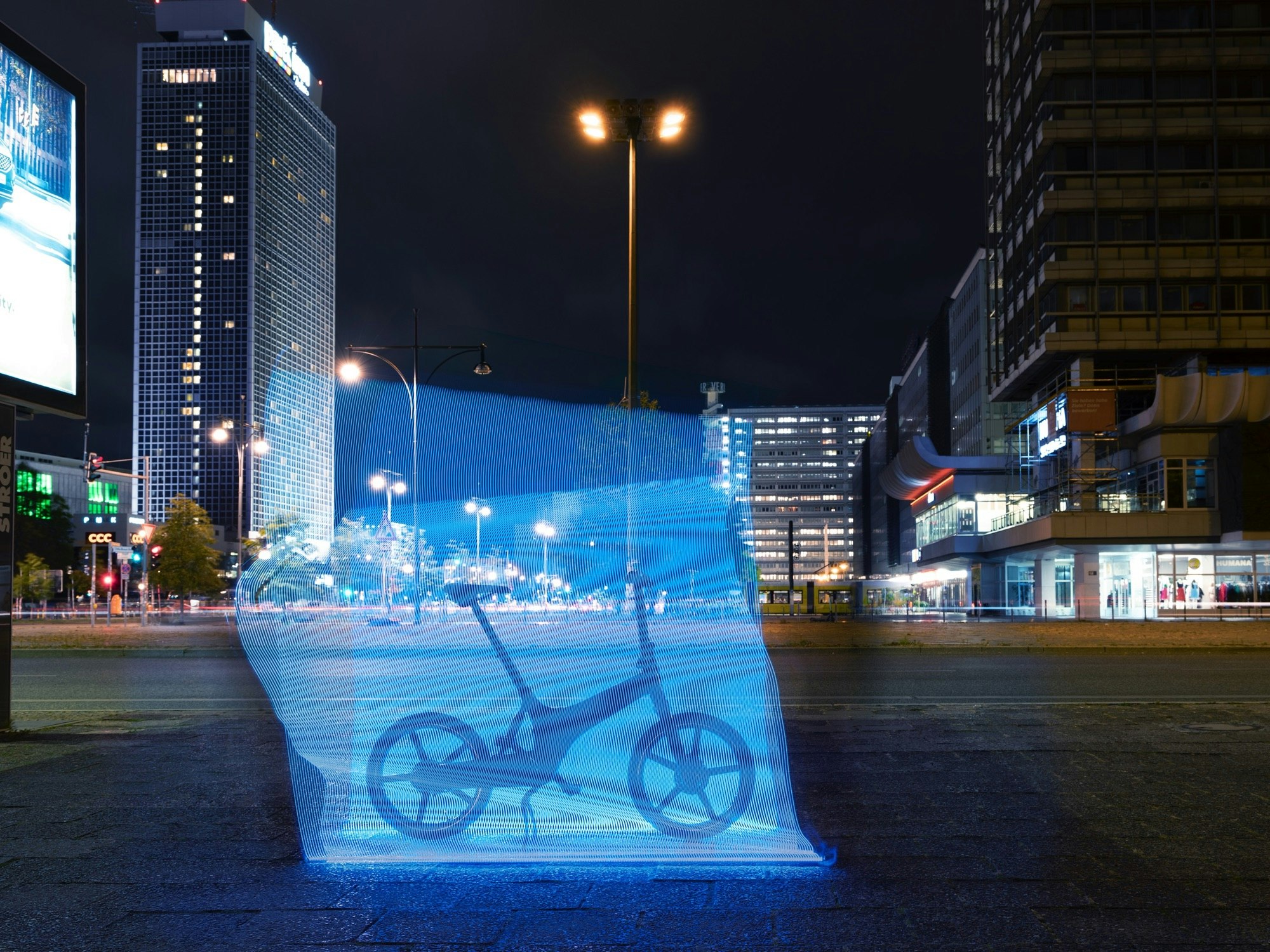 Long exposure photography of a bicycle on a city sidewalk at night, with blurred blue light, tall buildings, and street lights.