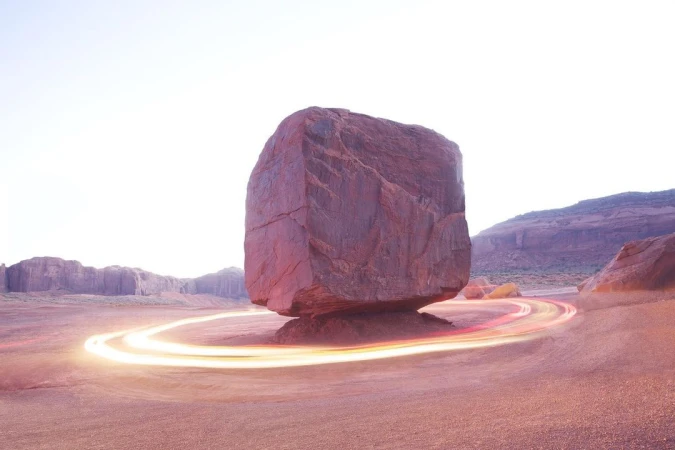Monument Valley landscape features a massive, cubed boulder precariously balanced on a small pedestal, encircled by light trails.