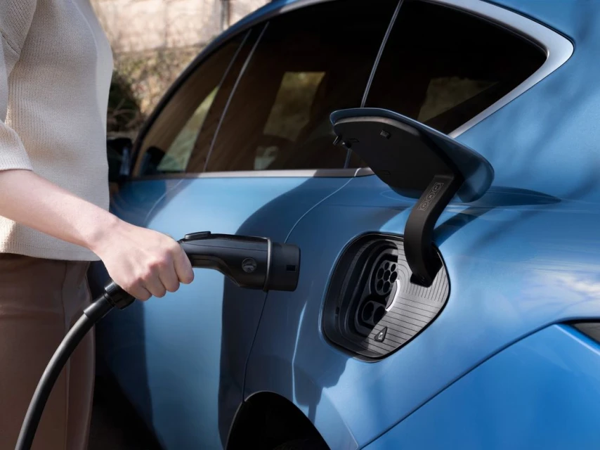 Person charges a light blue electric car with a black charging cable. The charging port is open and visible on the car's exterior.