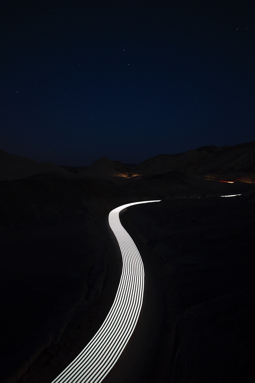 Long exposure shot of illuminated road winding through dark landscape under night sky with stars and minimal ambient light.