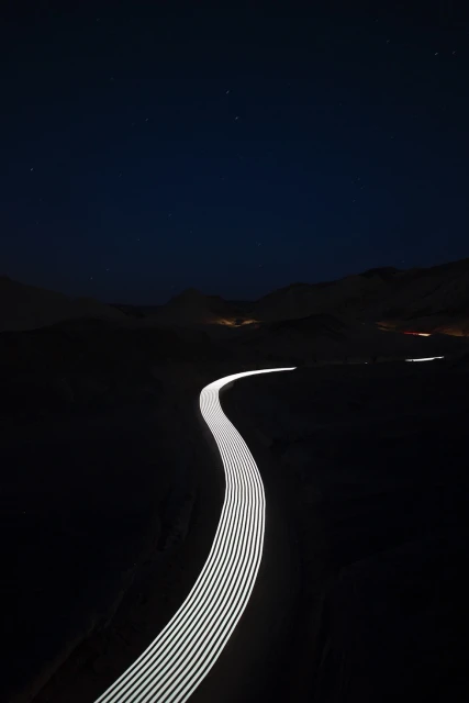 Long exposure shot of illuminated road winding through dark landscape under night sky with stars and minimal ambient light.