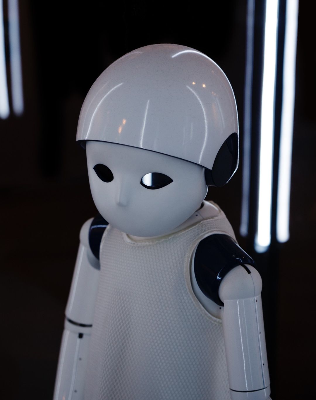 Close-up of a white humanoid robot with black accents, featuring a helmet-like head, set against a dark background with vertical lights.