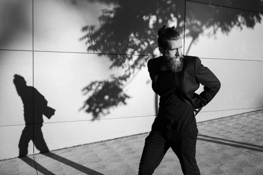 Black-and-white shot of a bearded man in a suit against a textured wall with tree shadows, in an artistic outdoor composition.