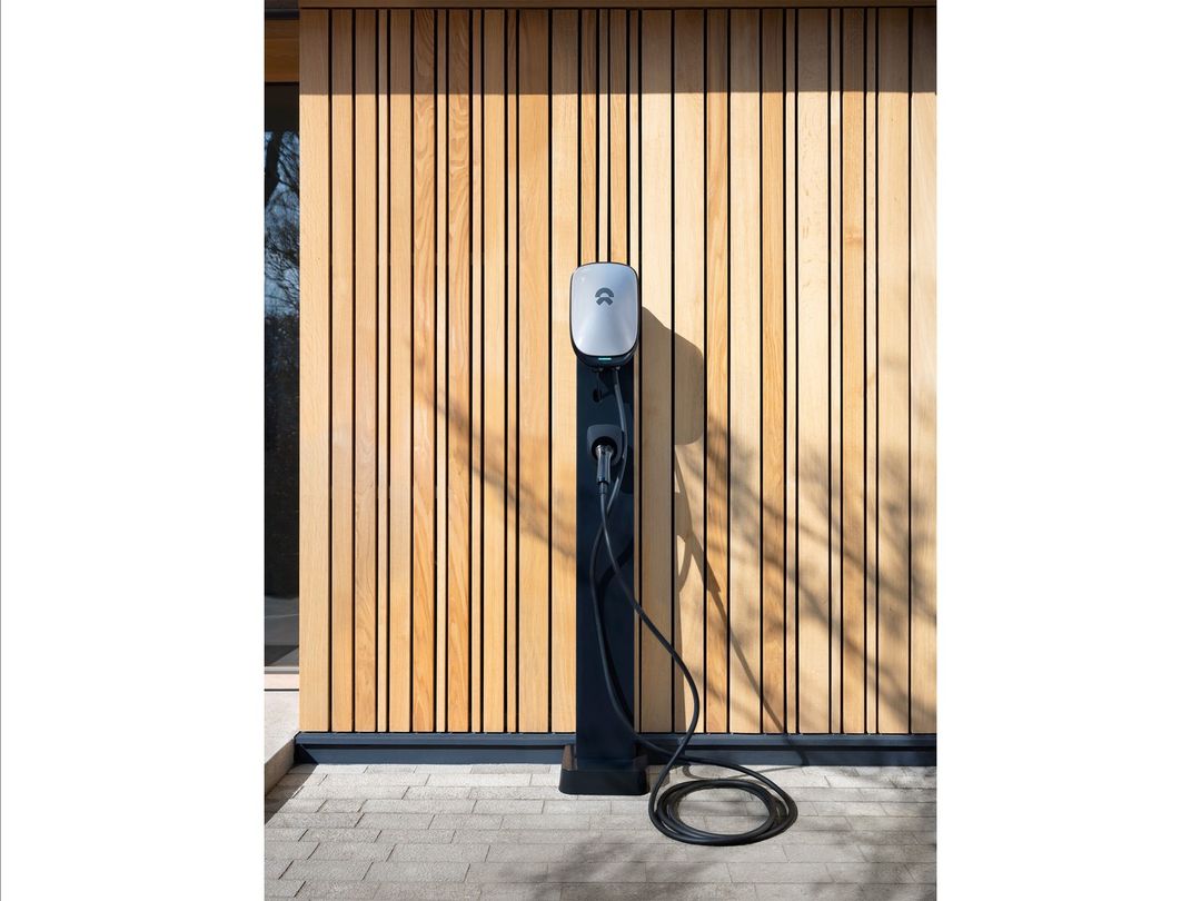 EV charger station on a black stand with a wood background outside on a sunny day. Charging cable is plugged into the EV charger.