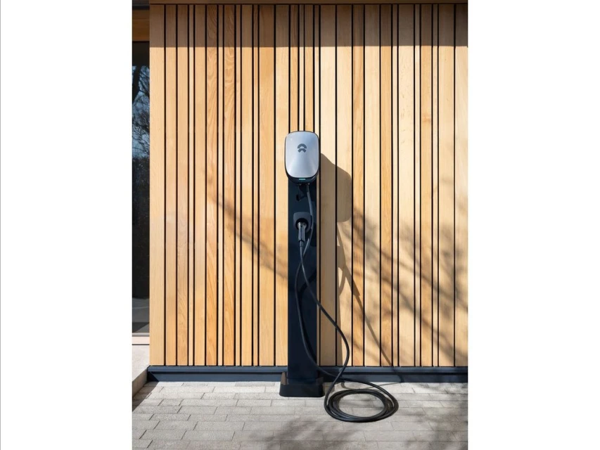 EV charger station on a black stand with a wood background outside on a sunny day. Charging cable is plugged into the EV charger.