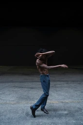 Shirtless man in blue trousers dancing outdoors on asphalt pavement under dark evening sky