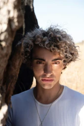 Portrait of a man with curly blonde hair and a white t shirt standing outdoors near a tree trunk.