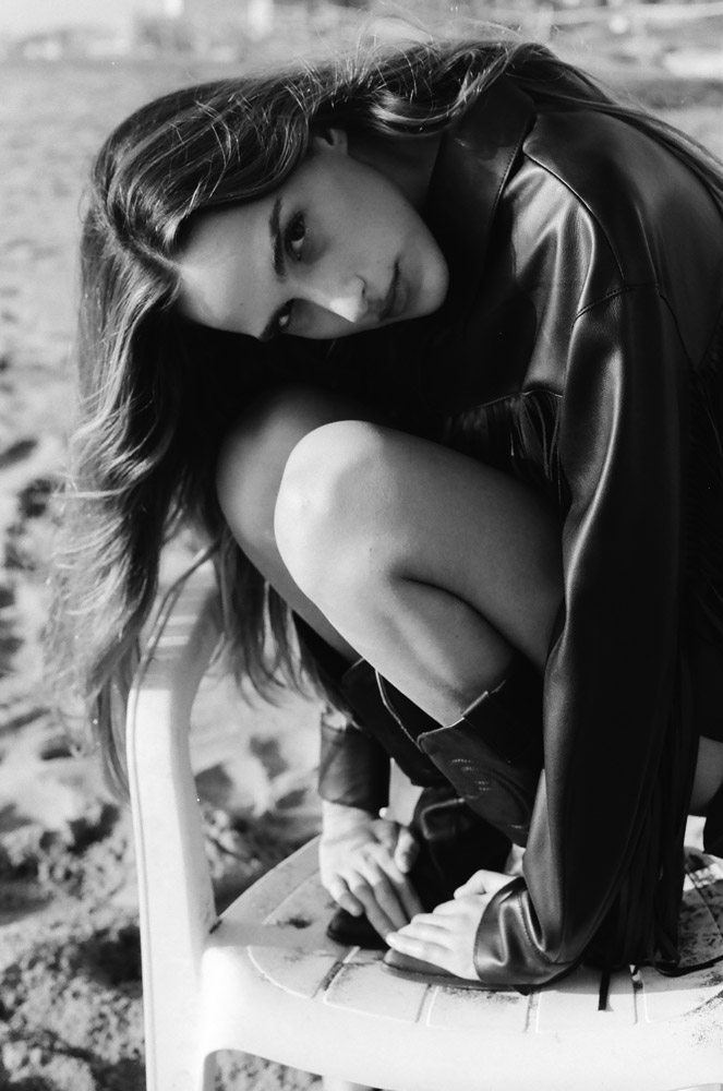 Black and white portrait of a woman in a leather jacket, crouching on a plastic chair on a beach, gazing intently at the camera.